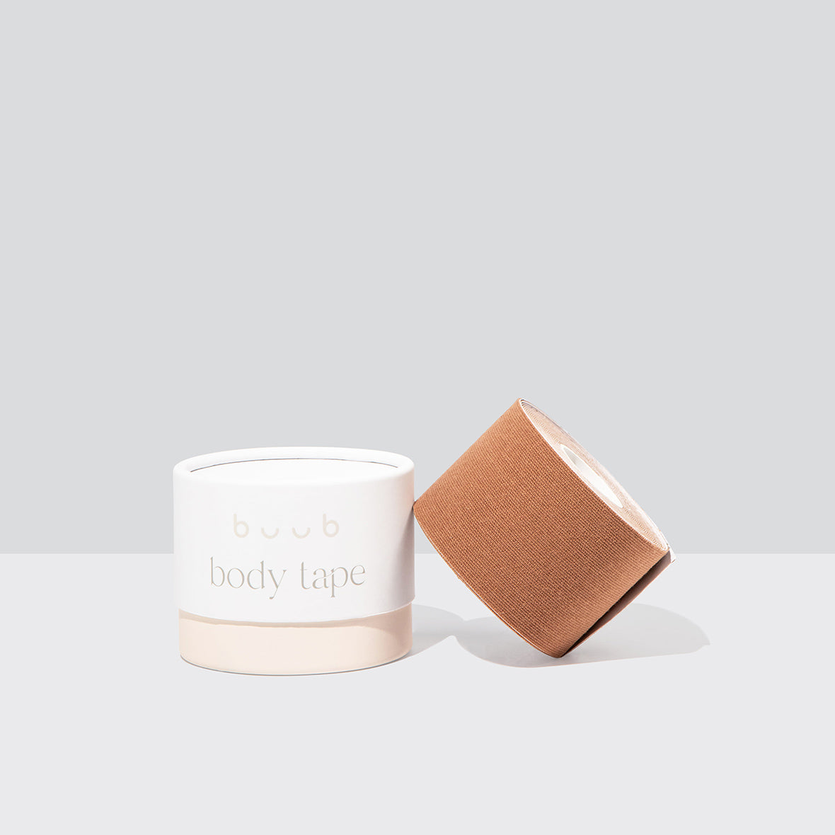 Studio product shot of Buub Body Tape in a tan shade. The roll of skin-friendly breast lift tape rests against its white and beige container on a light grey background.