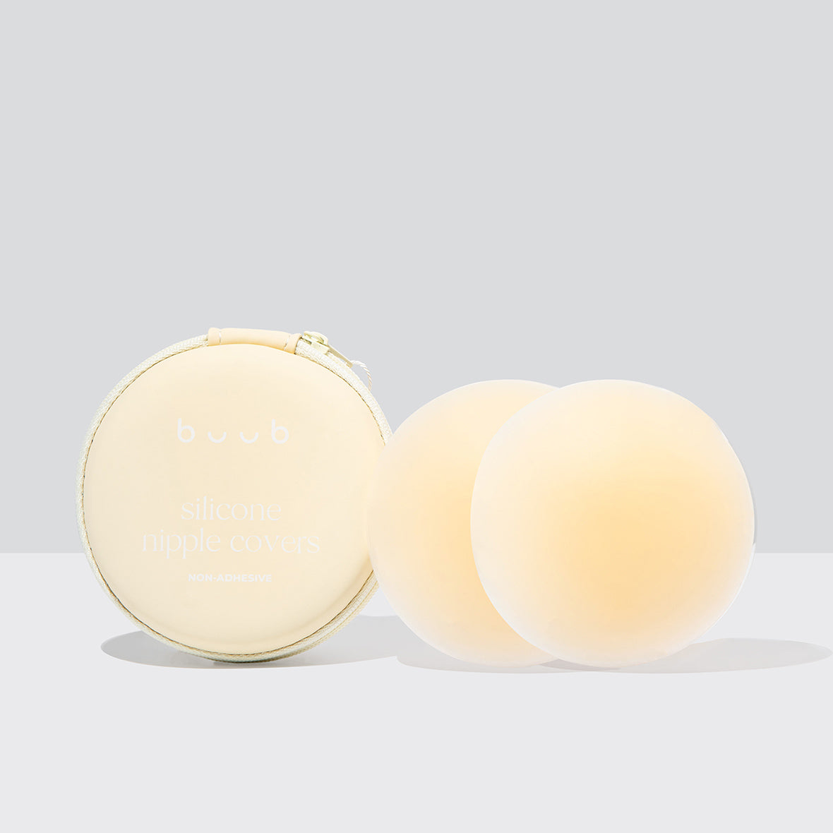 A pair of 8cm Buub non-adhesive silicone nipple covers in a light nude shade. The reusable seamless pasties are displayed next to their cream-coloured travel case.